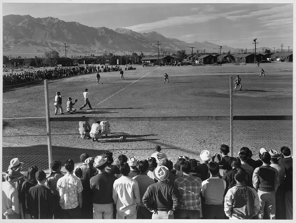 Spectators in the foreground watch a baseball game at Manzanar, with barracks and mountains beyond the field in Ansel Adams’s photograph.