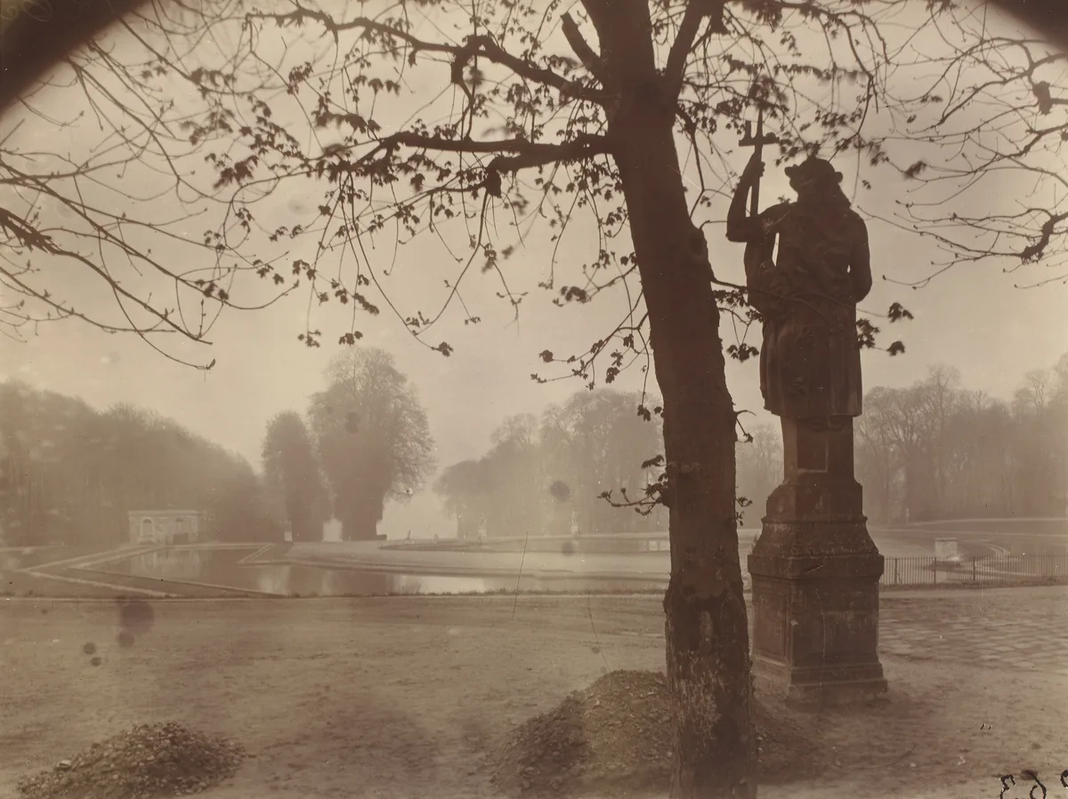 Atget’s Saint-Cloud photograph with a statue and dark tree at right, branches overhead and water opening toward distant trees.