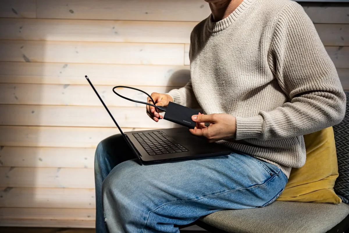 A person holding a portable Samsung drive and cable beside an open laptop