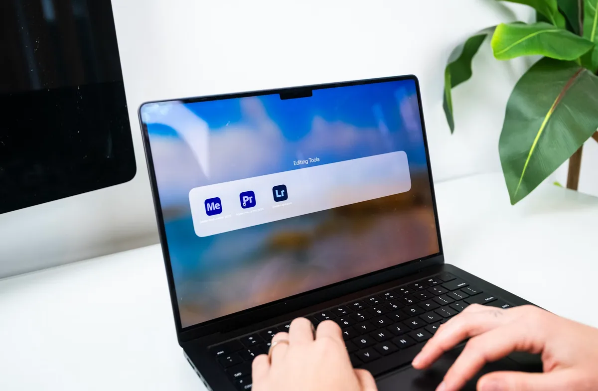 Hands using a MacBook displaying Lightroom, Premiere Pro and Media Encoder application icons