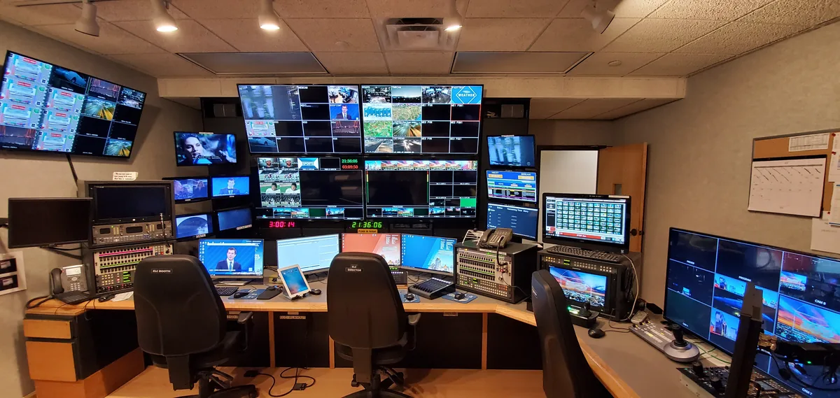 Television news production control room with desks, switching equipment and a wall of monitors.