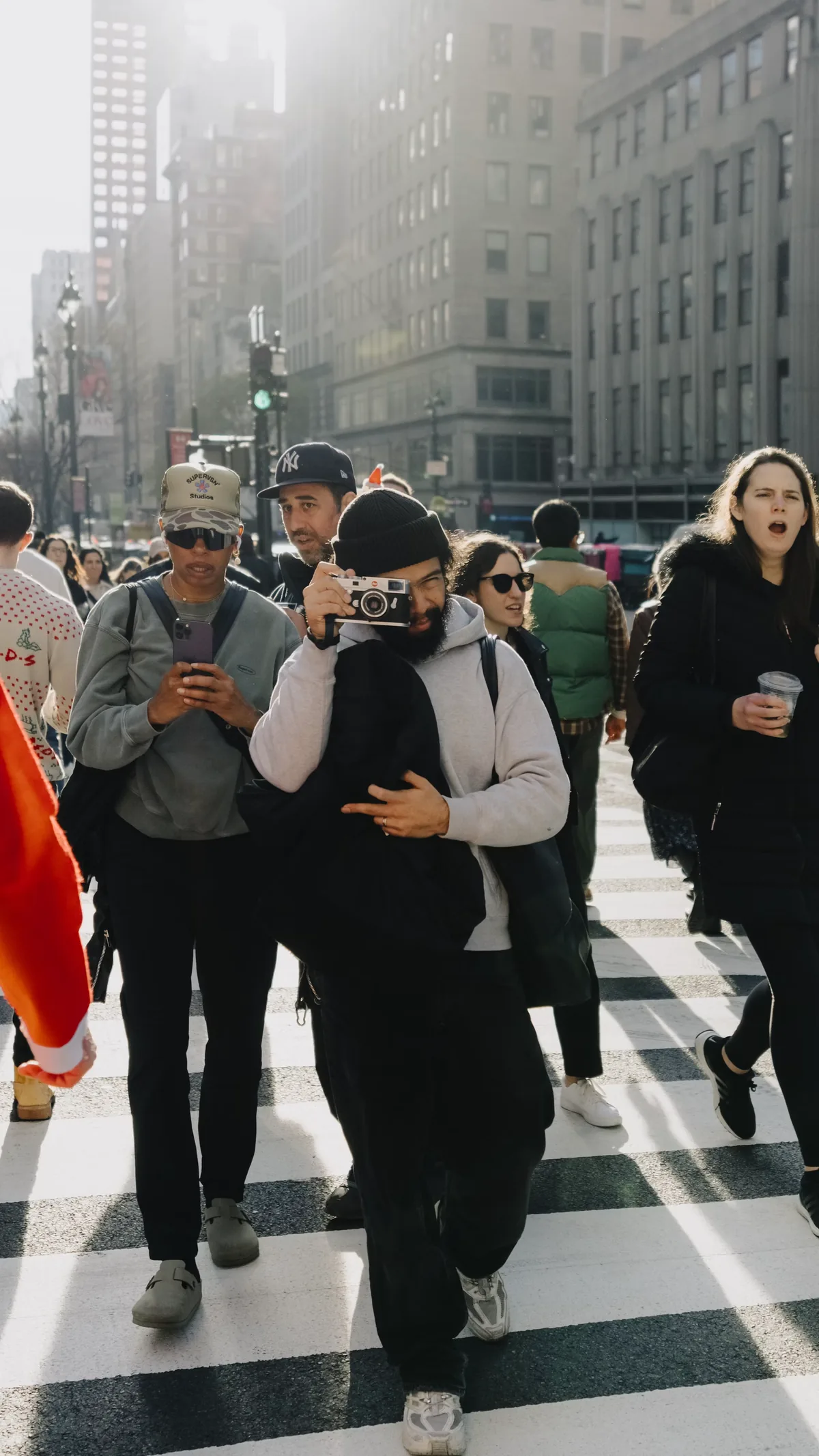 Pedestrians crossing a broad city street while one person raises a camera