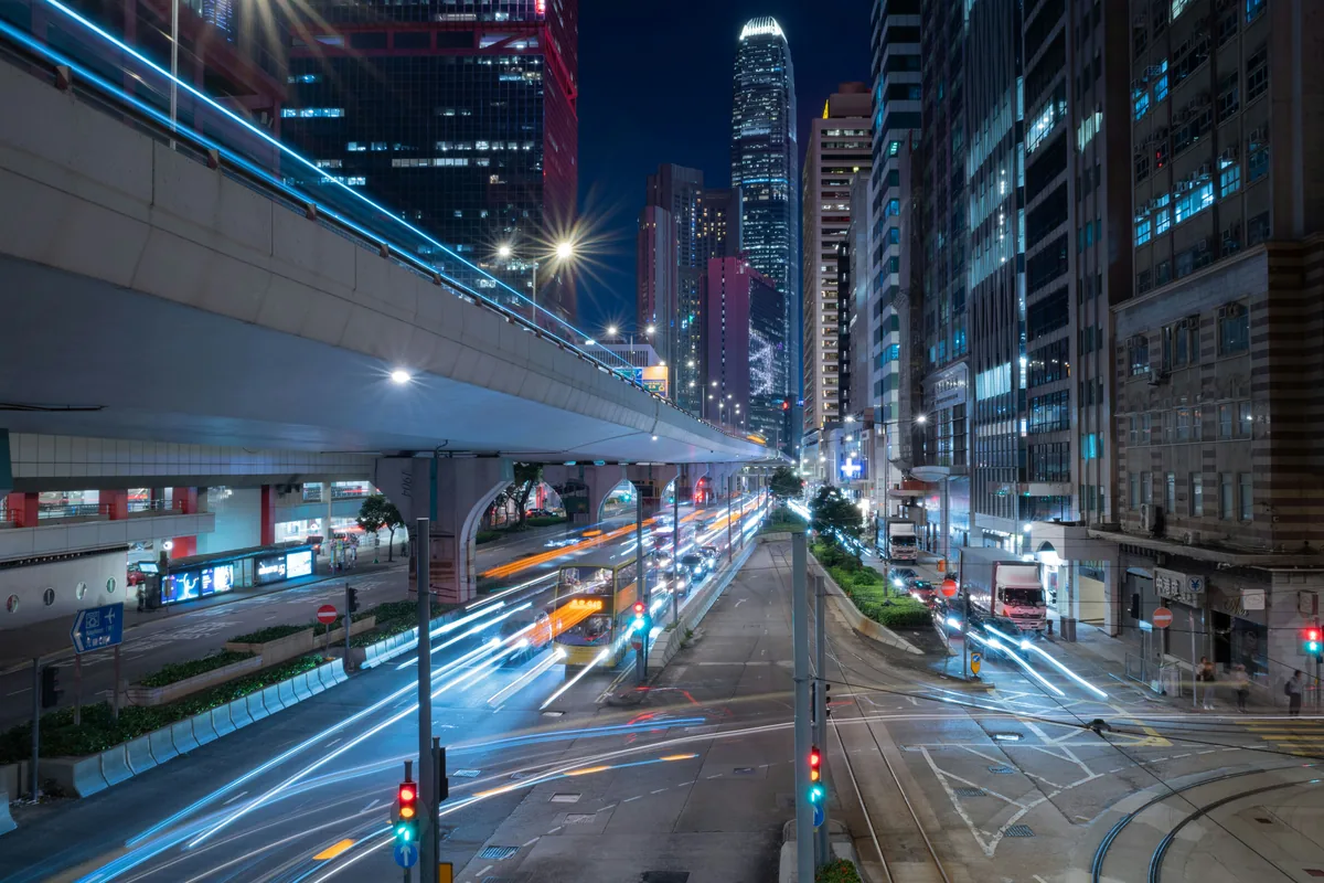 White, blue and orange vehicle light trails run between illuminated office towers beside an elevated road at night.