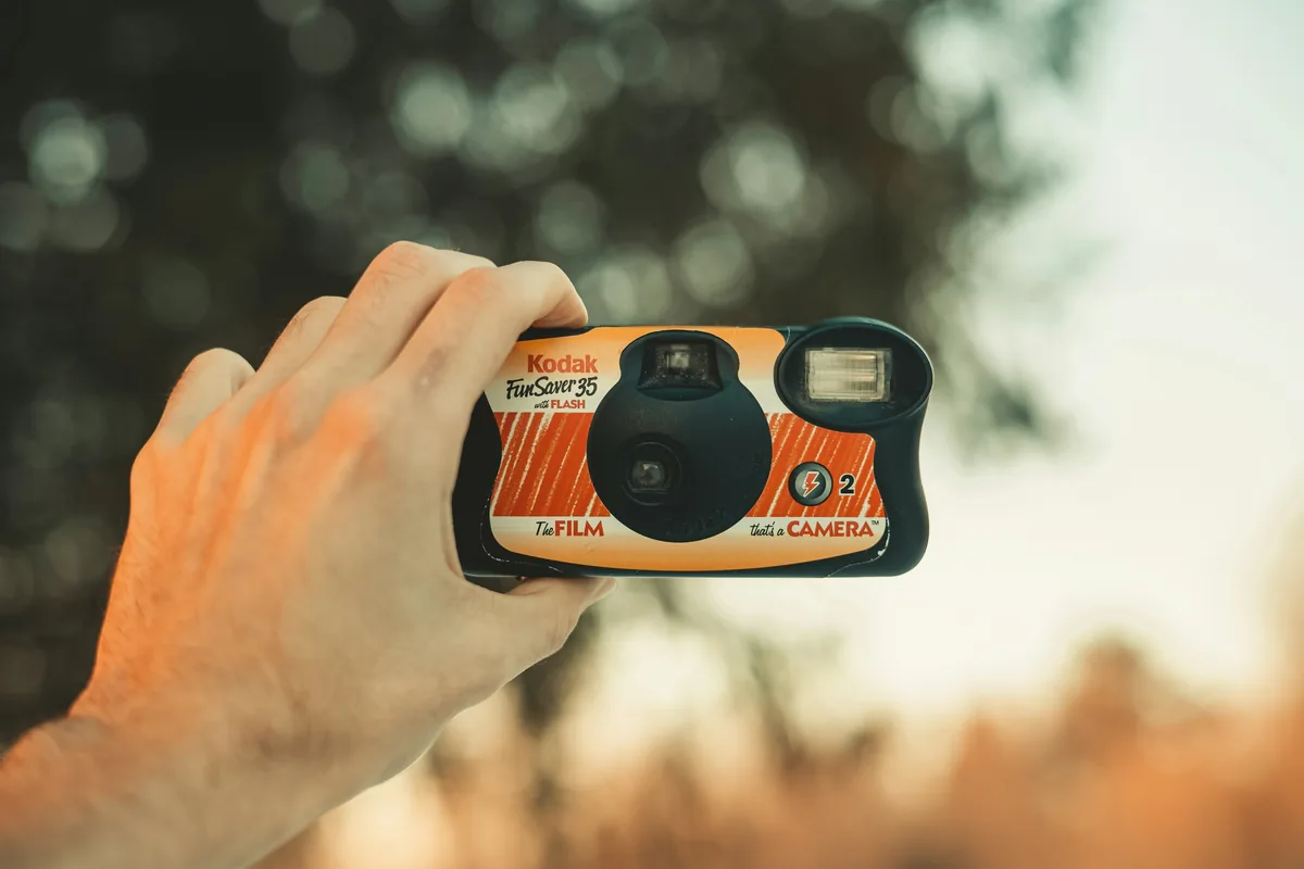 A hand holds a Kodak FunSaver 35 single-use camera with a built-in flash outdoors.