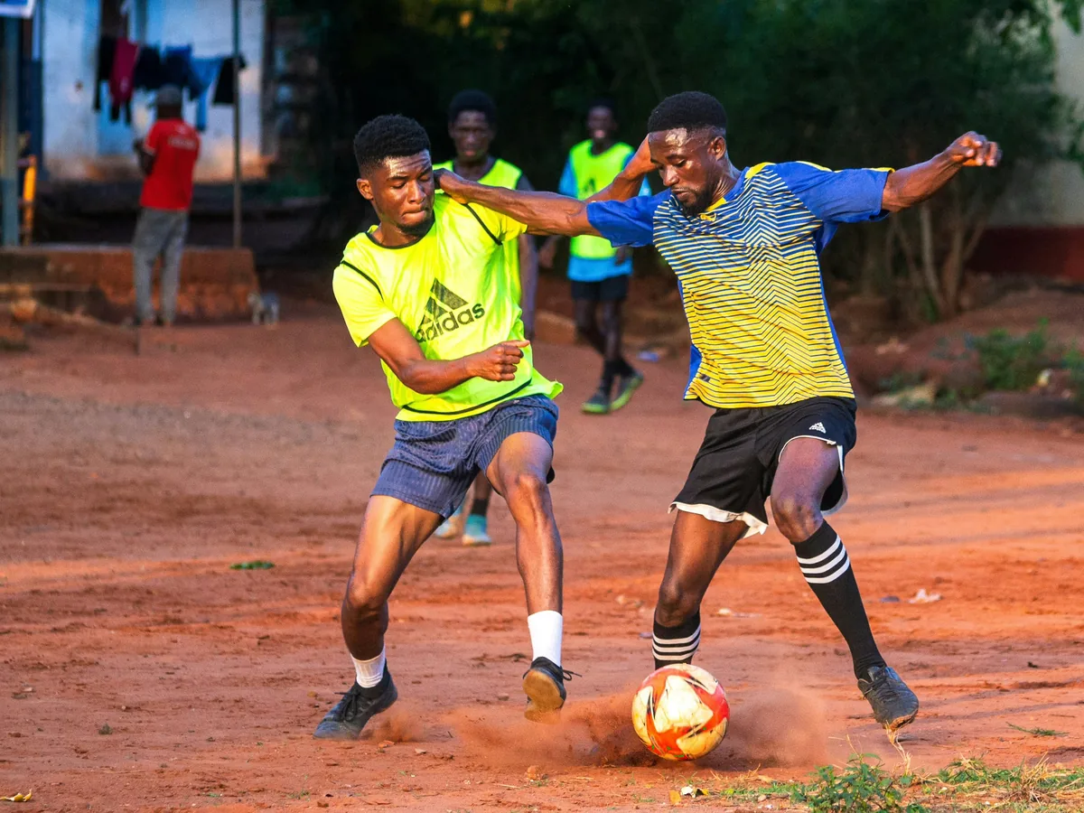 Two football players contesting a ball on a dirt pitch