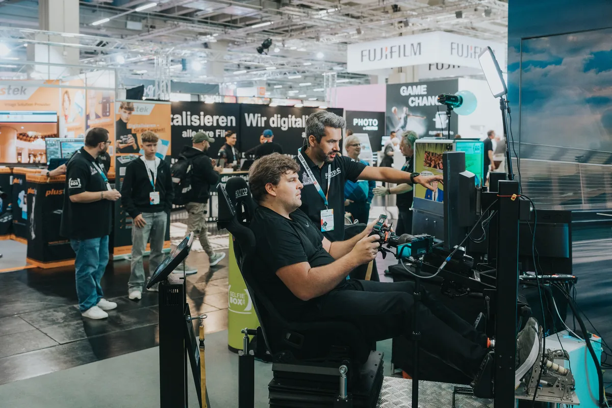Visitor trying a seated simulator while an exhibitor points at its display at Imaging World 2025.