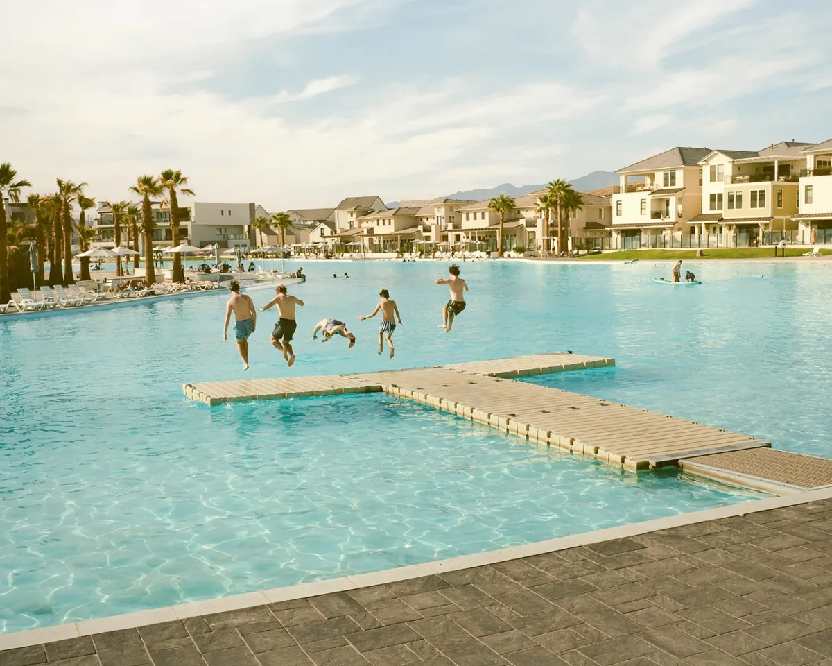 Children jumping from a floating platform into blue water beside houses and palm trees.