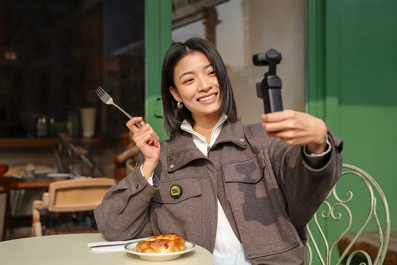 Woman wearing an Insta360 Mic transmitter while holding a small gimbal camera at an outdoor cafe.