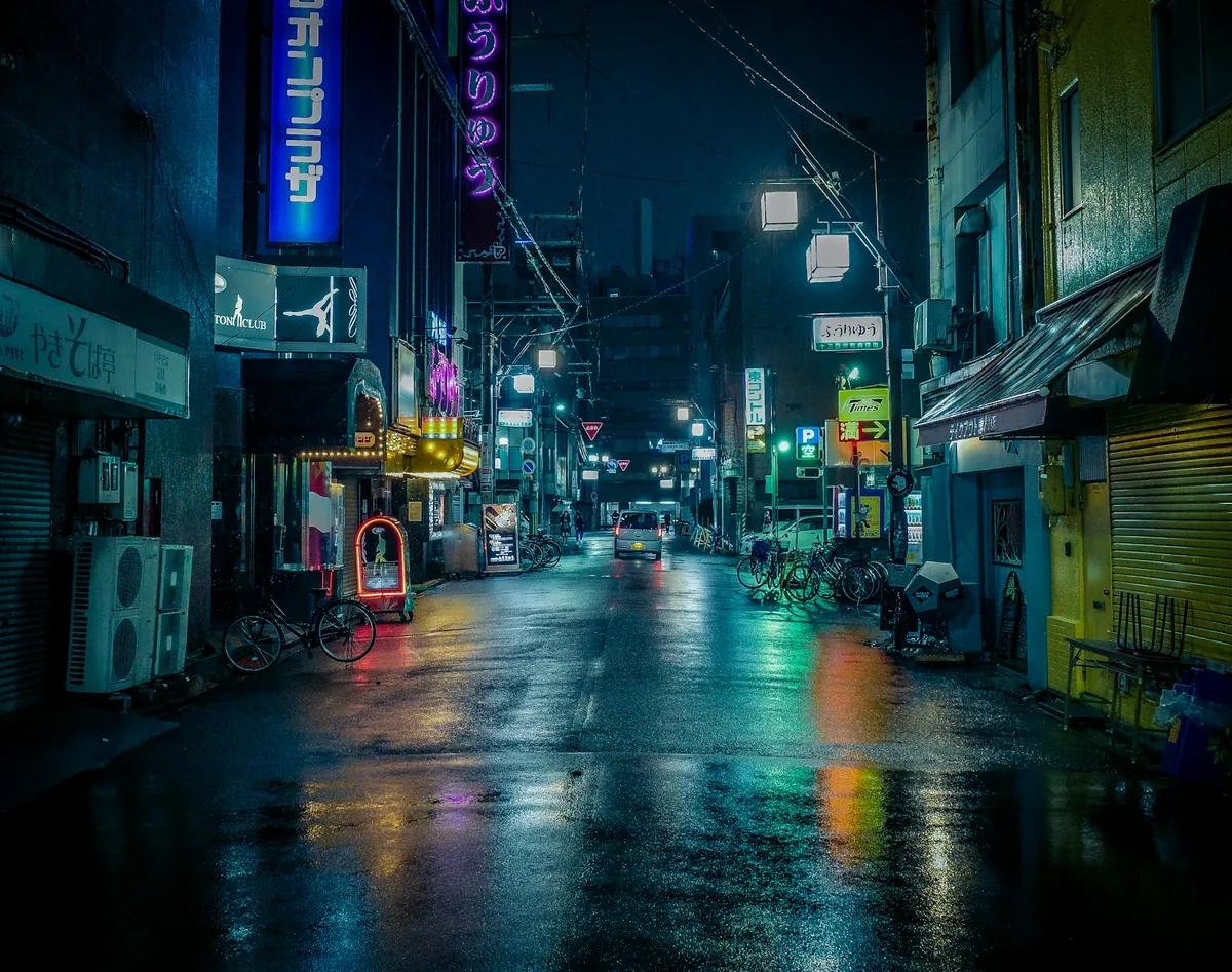 Blue, green and red signs reflected on a wet Japanese street at night, with a car in the distance.