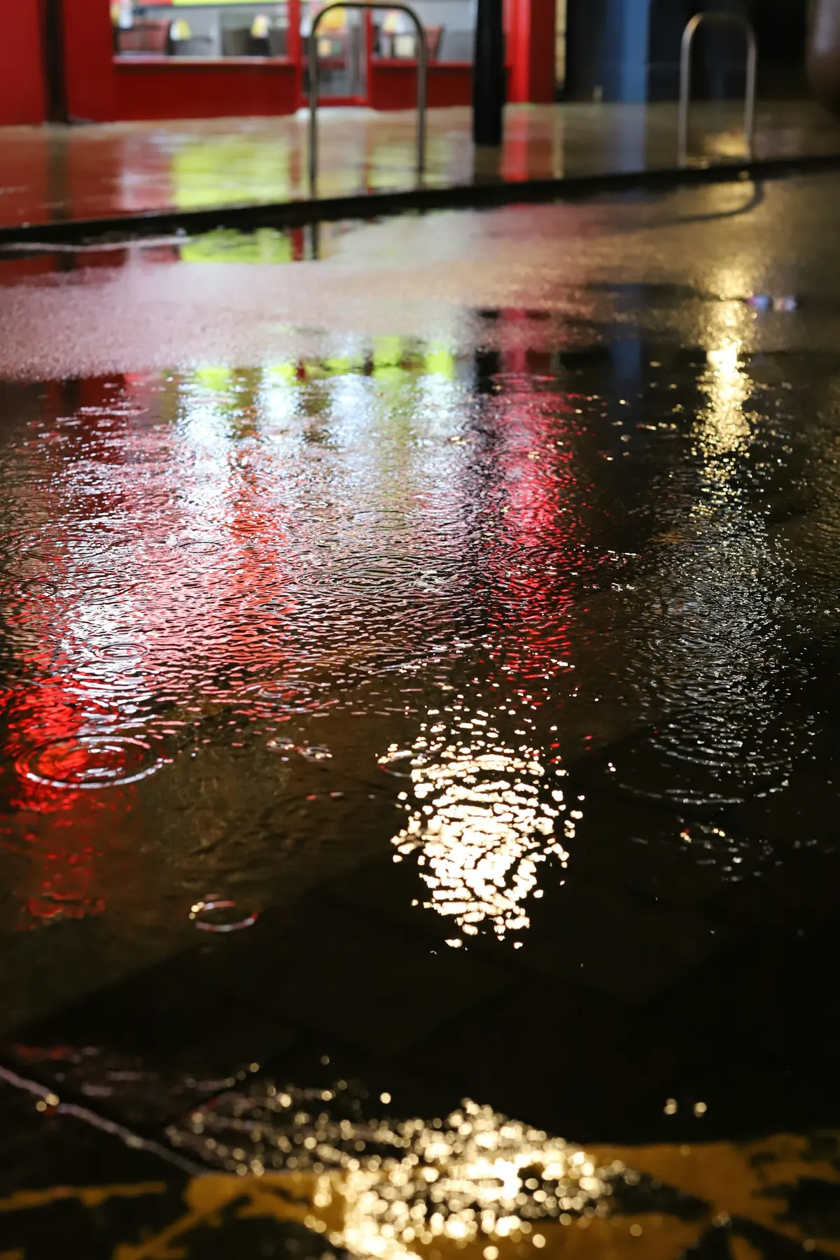 Red, yellow and white storefront lights reflected in rippling water on a wet street.