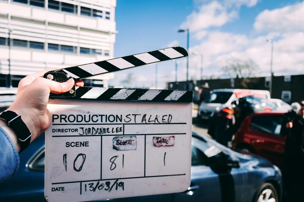 A hand holding an open black-and-white clapperboard outdoors beside parked vehicles.