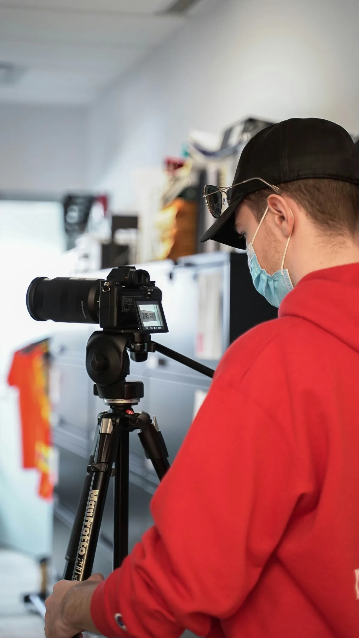 Operator in a red shirt and face mask using a camera on a tripod indoors