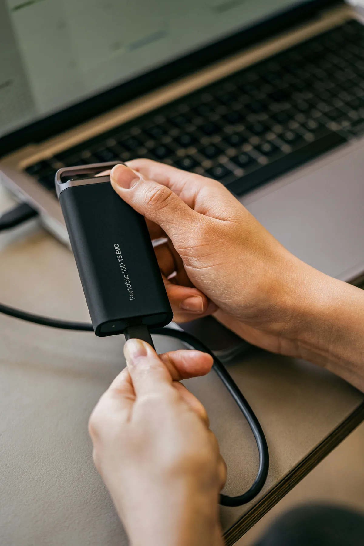 Hands holding a portable SSD and cable in front of a laptop.