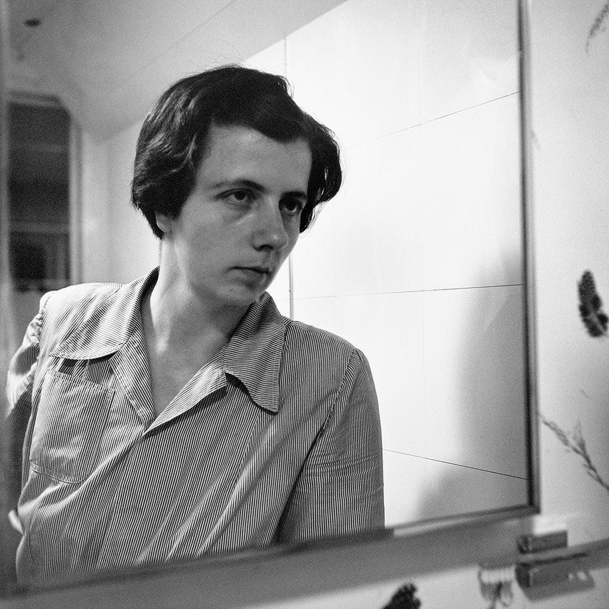 Vivian Maier reflected in a mirror, wearing a striped shirt beside a tiled wall